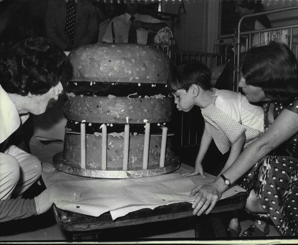 5 year old Peter of Wollongong Patient in the William Stewart ward of the Royal Alexandra Hospital for Children Camperdown, cutting into the 185 lb Hamberger shaped birthday cake with the help of Miss J. Noble director of Nursing and Ronald McDonald. McDo