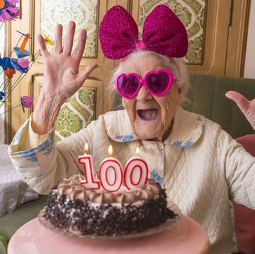 funny birthday with 100 years old birthday cake, elderly woman wearing pink heart sunglasses, huge pink bow headband, smiling big