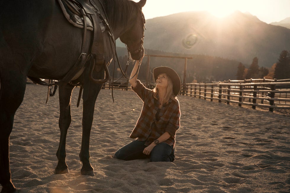a person kneeling in a sandy arena beside a horse during sunset