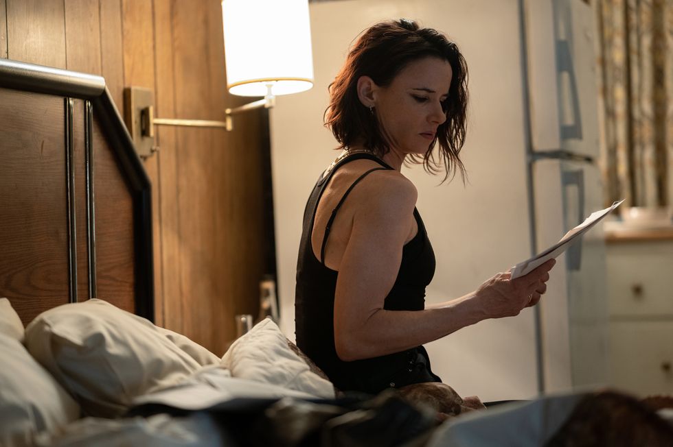 juliette lewis sits on a bed in a motel in yellowjackets wearing a black tank top and looking at a piece of paper