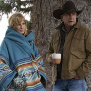 beth wearing a blue oversized jacket with a western design and hood she standing in front of a tree with john who is wearing a brown jacket, cowboy hat and holding a coffee cup