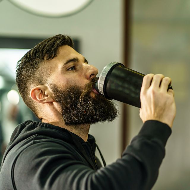 young adult man with beard drinking bottle supplement shake in training at gym side view copy space real people
