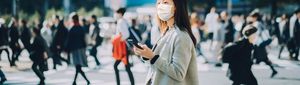 young asian businesswoman commuting in busy downtown city street with protective face mask and using smartphone to protect and prevent from the spread of viruses during covid 19 health crisis