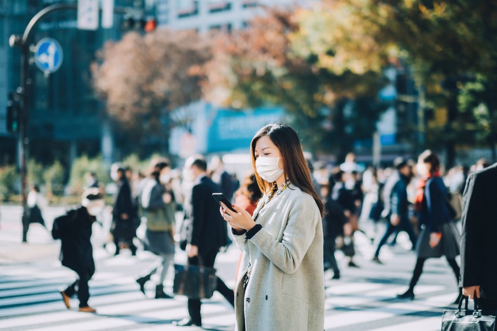 young asian businesswoman commuting in busy downtown city street with protective face mask and using smartphone to protect and prevent from the spread of viruses during covid 19 health crisis