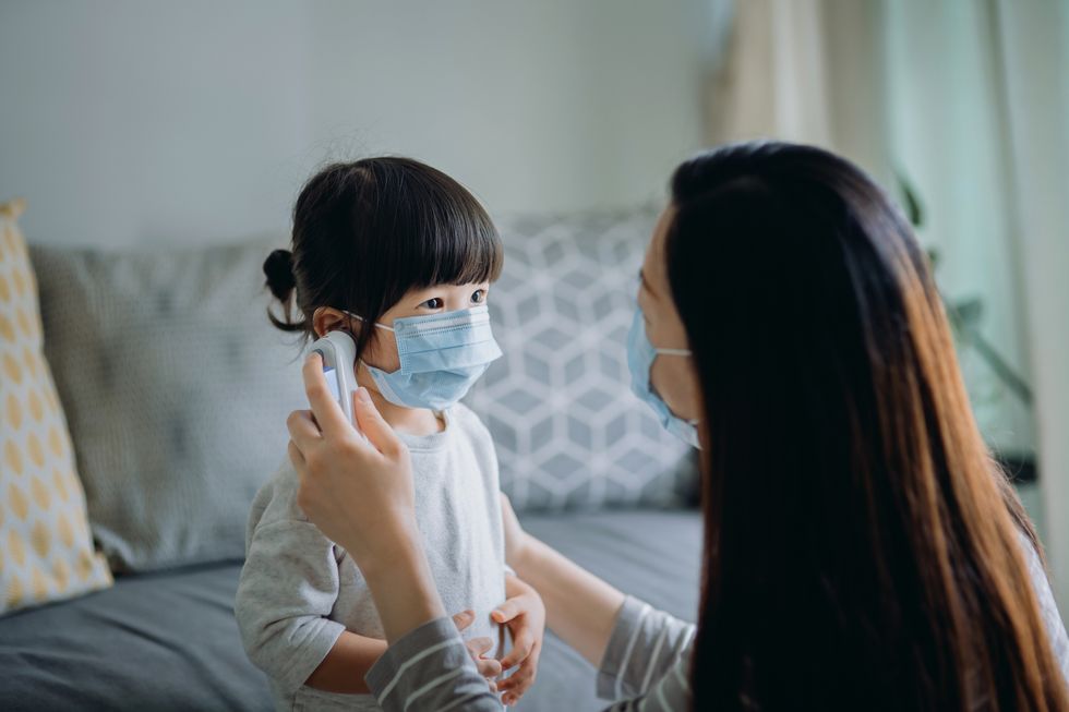 young asian mother checking sick daughter's temperature with a digital thermometer in the living room, both of them are wearing a surgical mask to prevent the spread of cold and flu and viruses