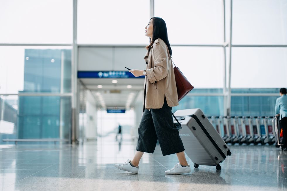 young asian woman carrying suitcase and holding smartphone on hand, walking in airport terminal ready to travel travel and vacation concept business person on business trip