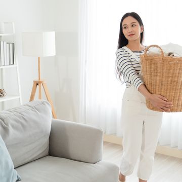 young asian woman happy taking messy dirty clothes into the basket in living room at home. beautiful cleaning service worker cleaning and housekeeping. housework and chores concept