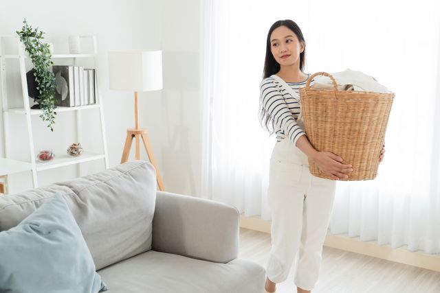 young asian woman happy taking messy dirty clothes into the basket in living room at home. beautiful cleaning service worker cleaning and housekeeping. housework and chores concept