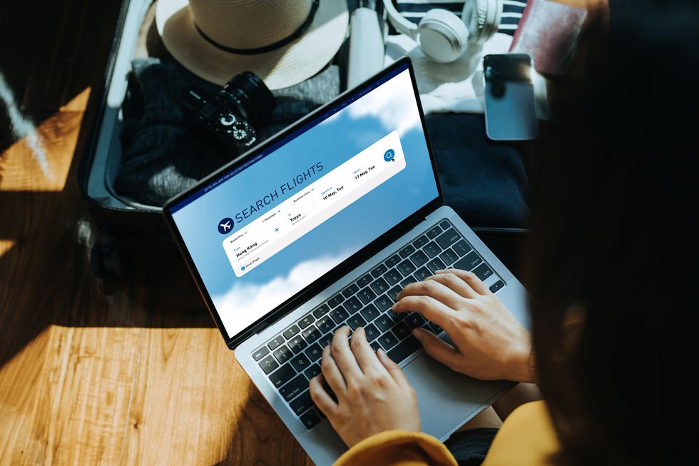 young asian woman shopping online for flight ticket on airline website with laptop at home, sitting next to suitcase with sun hat, camera, clothings, headphones, smartphone and passport getting ready for vacation travel and vacation concept