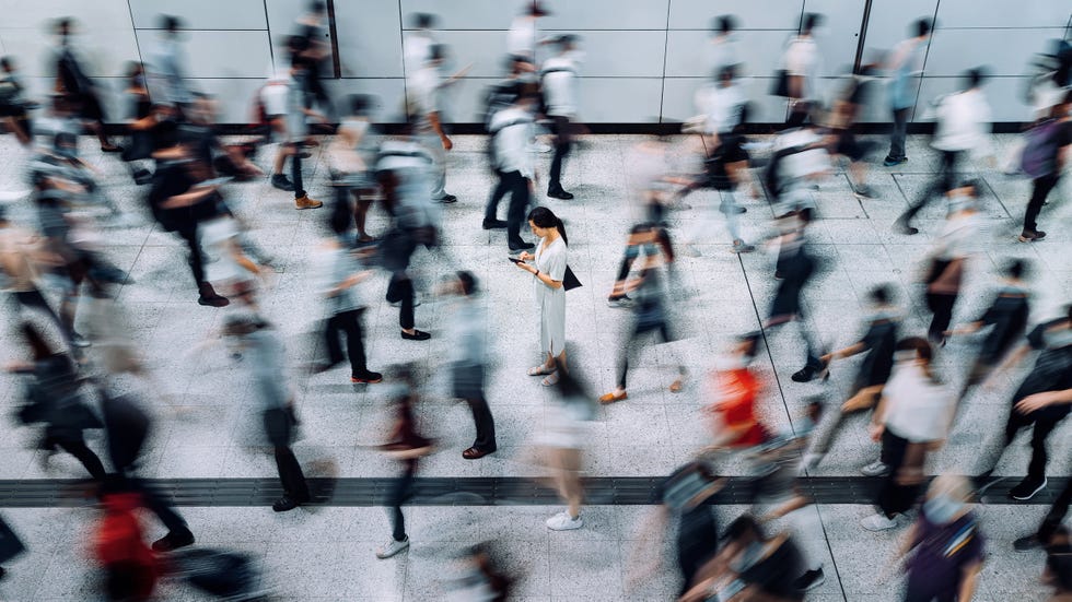 young asian woman using smartphone surrounded by commuters rushing by in subway station during office peak hours in the city