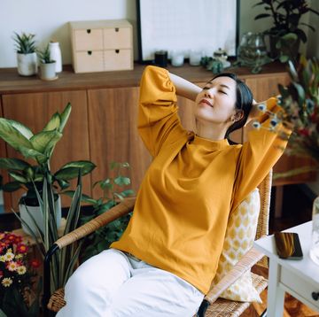young asian woman with hands behind her head, taking a break and relaxing on armchair at cozy home, surrounded by green houseplants. lifestyle, wellbeing and wellness concept