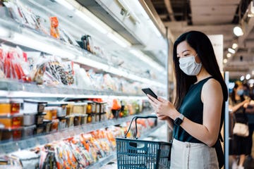 young asian woman with protective face mask holding shopping basket and using smartphone while grocery shopping in a supermarket