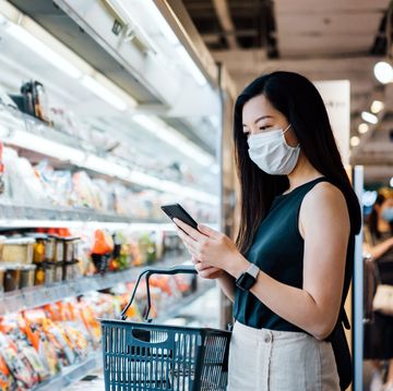 young asian woman with protective face mask holding shopping basket and using smartphone while grocery shopping in a supermarket