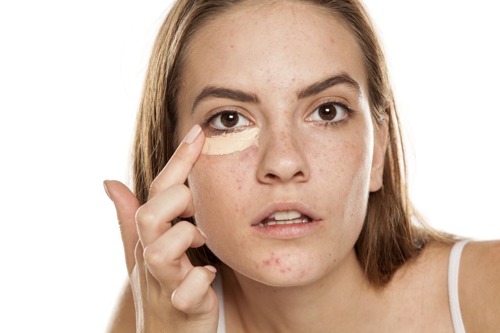 young beautiful girl applying concealer with fingers under her eyes on white backgeound