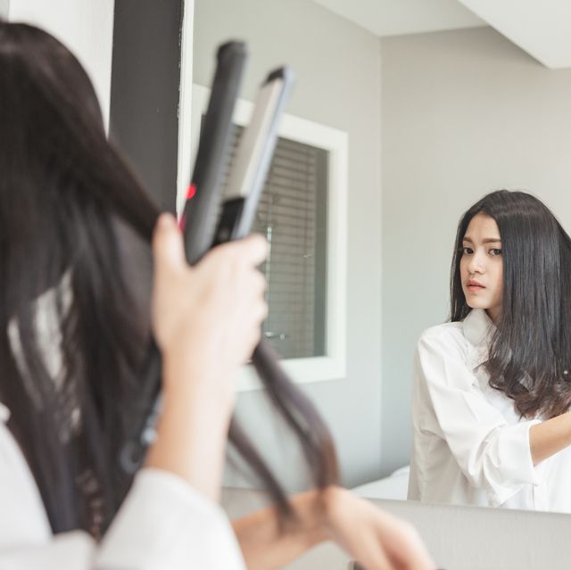 young beautiful woman getting dressed and using hair straighteners in front of mirror in room