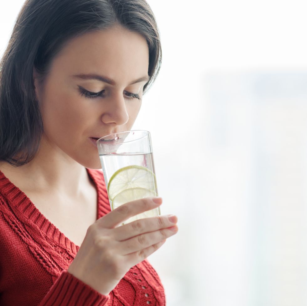 young beautiful woman in red sweater with glass of water with lime, woman stands near window in skyscraper on cloudy day healthy drink, natural antioxidant, vitamins in urban life