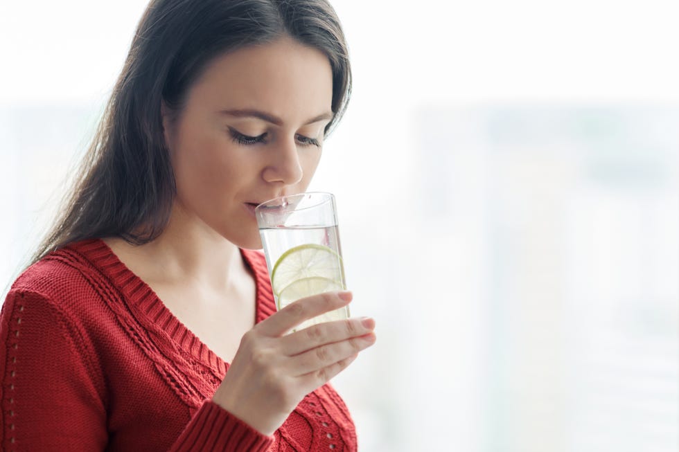 young beautiful woman in red sweater with glass of water with lime, woman stands near window in skyscraper on cloudy day healthy drink, natural antioxidant, vitamins in urban life