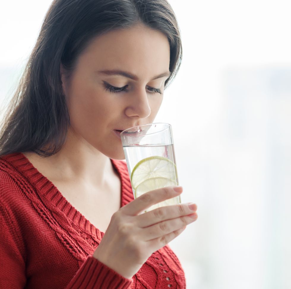 young beautiful woman in red sweater with glass of water with lime, woman stands near window in skyscraper on cloudy day healthy drink, natural antioxidant, vitamins in urban life