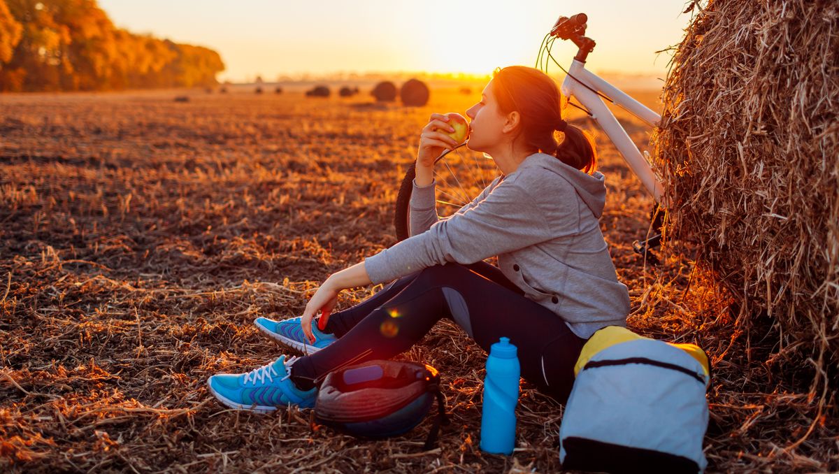 Young bicyclist having rest after a ride in autumn field at sunset. Woman eating by haystack