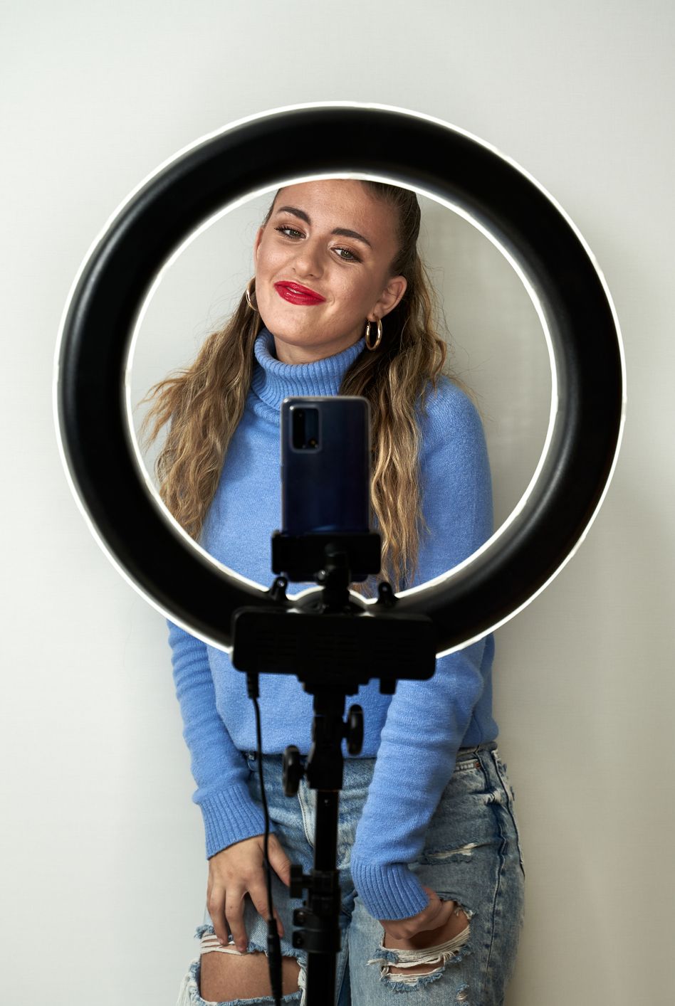 young blonde caucasian teenager influencer recording a video with smart phone in ring light on white background