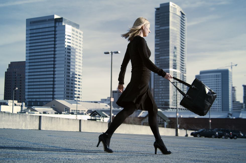 A young business woman walks across a parking lot with high-rises in the background.