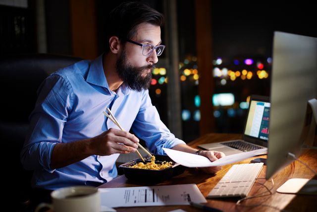 young businessman looking at computer and eating takeaway at office desk at night