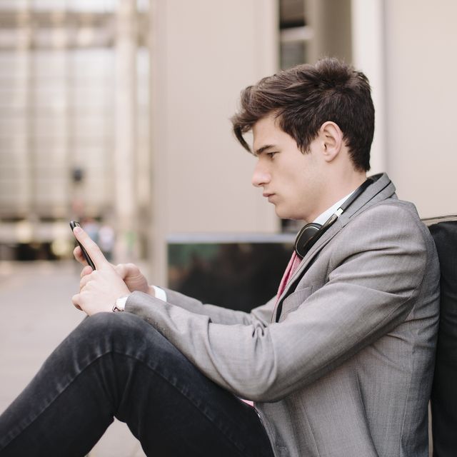 Young city businessman sitting on sidewalk reading smartphone messages