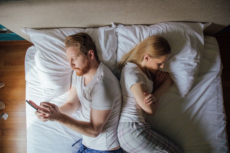 Young couple lying in bed back to back, man using phone and woman frowning