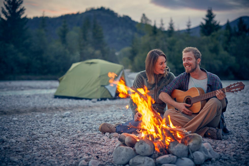young couple sitting by campfire playing guitar