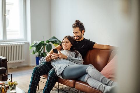 smiling young woman using mobile phone while reclining on boyfriend sitting on sofa at home