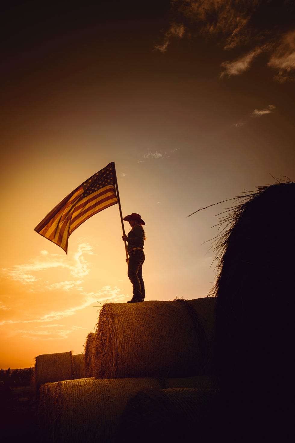 Young Cowgirl Stands On Top Of A Huge Pile Of Hay Bales At Sunset Holding The American Flag