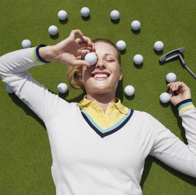 young female golfer lying on green, holding ball in front of eye, view from above