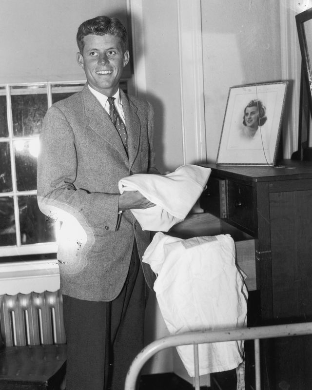 john f kennedy stands next to a dresser with an open drawer and holds a folded sheet, he smiles and wears a suit and tie