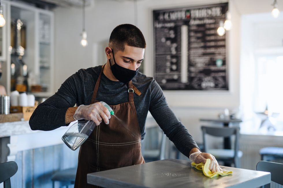 young man barista with face mask and gloves standing in coffee shop, disinfecting tables