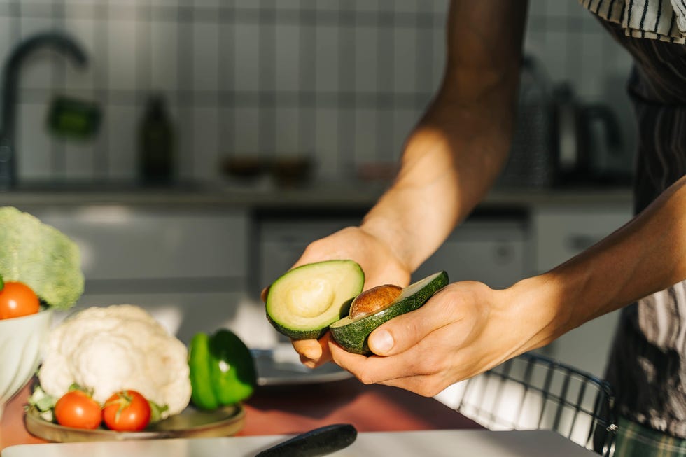 hands holding a halved avocado with visible flesh and seed surrounded by fresh vegetables