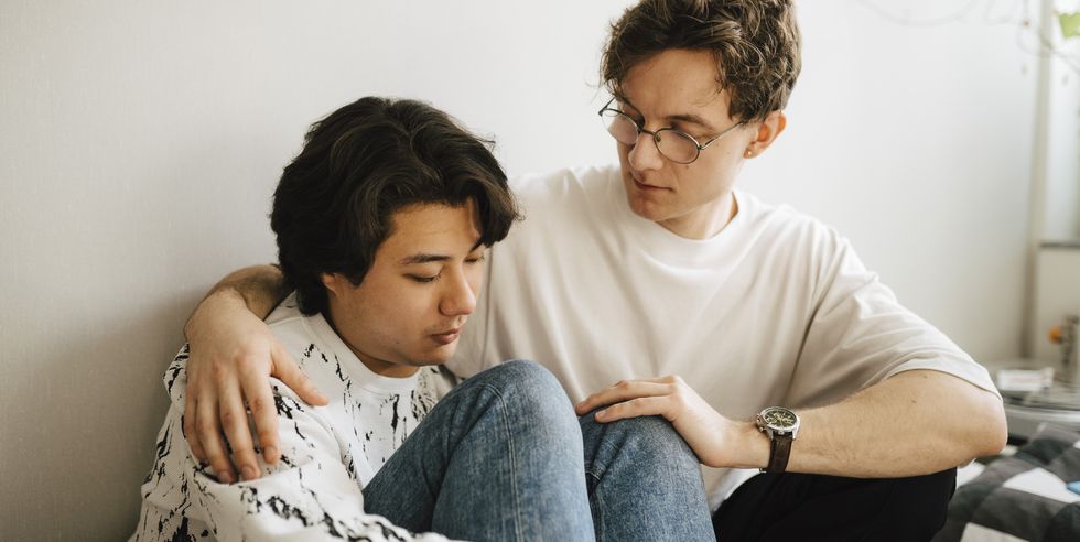 young man sitting by worried male friend in bedroom
