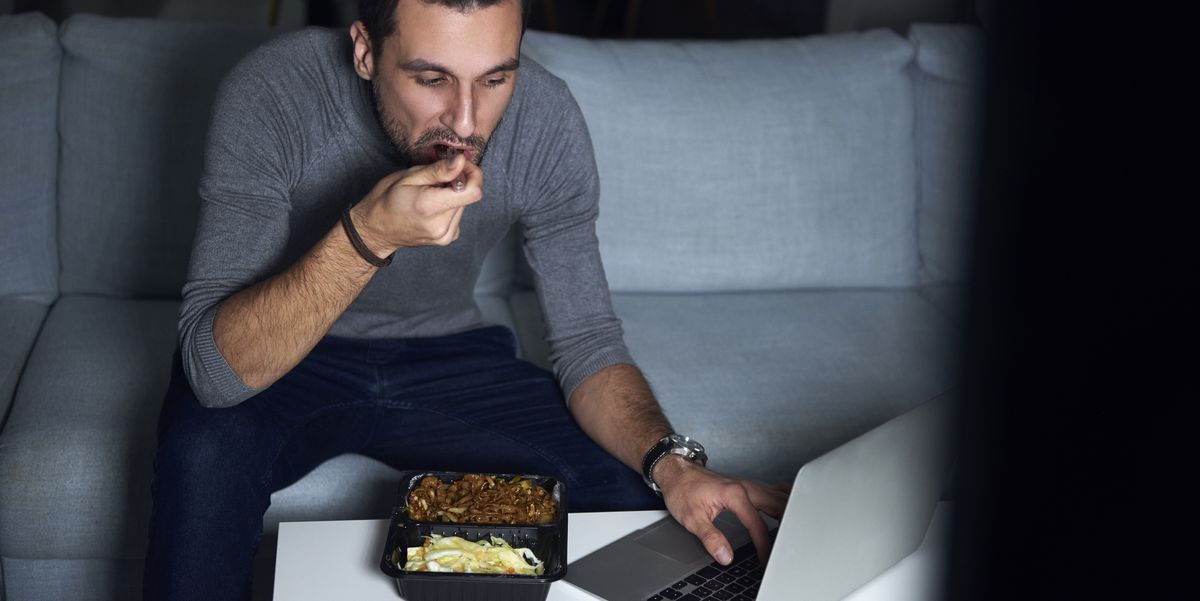 Young man sitting on sofa in evening eating takeaway and using laptop