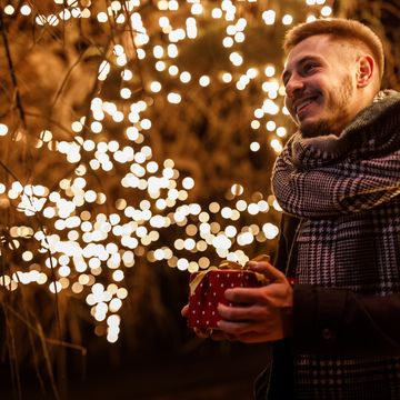 young man standing by the glistening christmas lights at the park, holding a christmas gift box