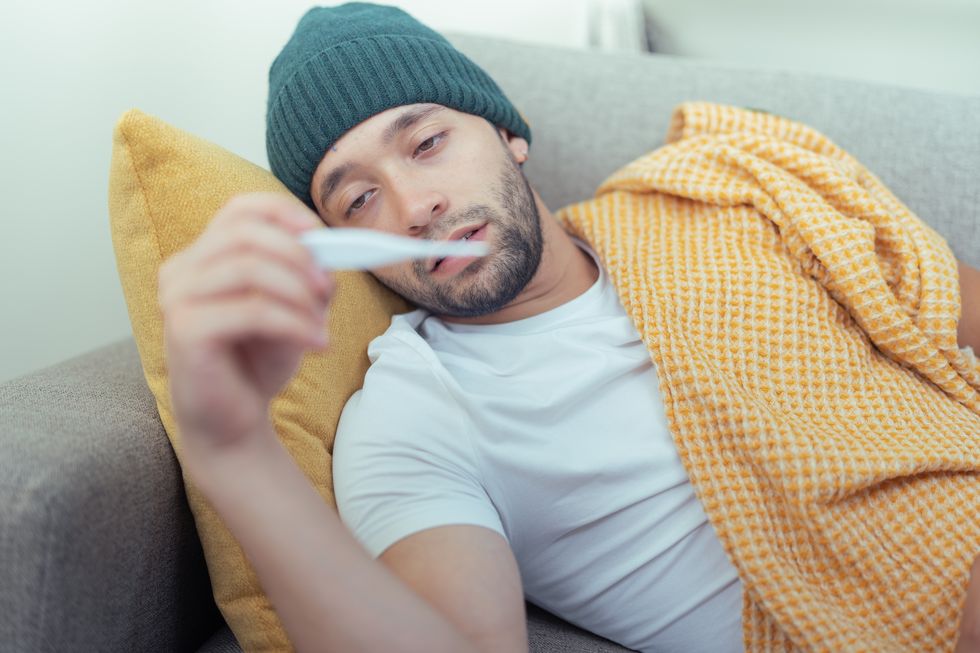 young man with fever taking his temperature, lying on the living room couch