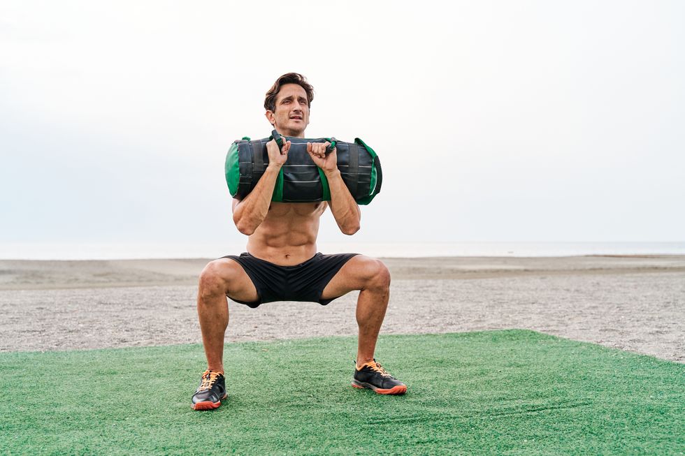young muscular man training on the beach shirtless doing a front squat lifting a sandbag