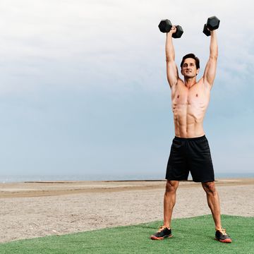 young muscular man training shoulder lifts on the beach shirtless lifting dumbbells