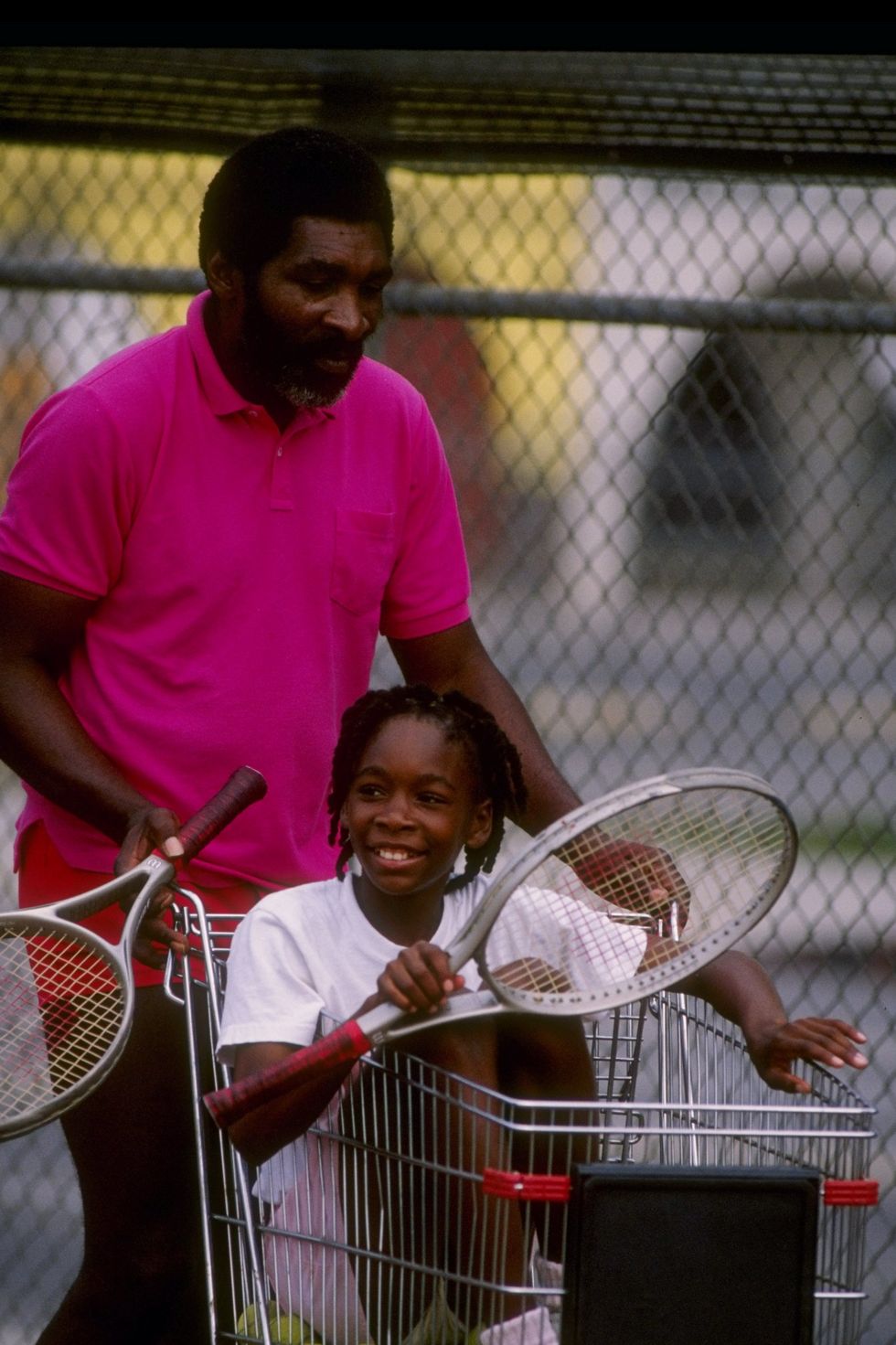a man holds a tennis racket and pushes a shopping cart that a young girl sits in, below here are many tennis balls and she also holds a tennis racket as she smiles and looks left
