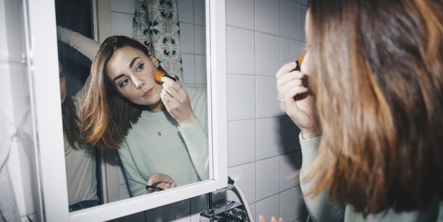 Young woman applying blush looking in mirror at college dorm bathroom
