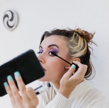 young woman applying mascara looking in smart phone while standing against white wall
