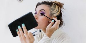 young woman applying mascara looking in smart phone while standing against white wall