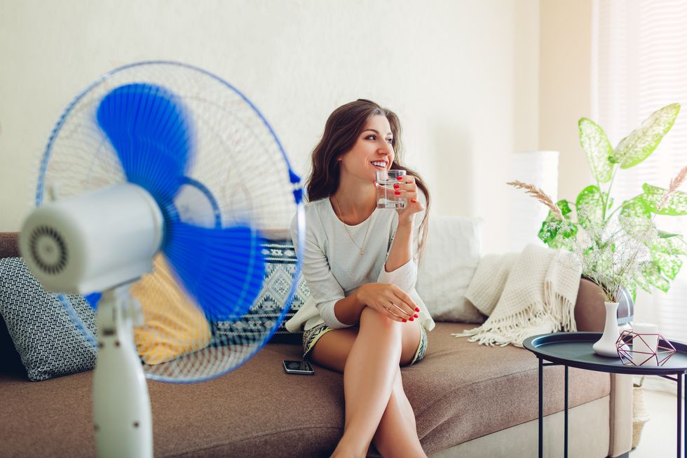 young woman cooling down by ventilator at home while drinking water and hanging in phone summer heat