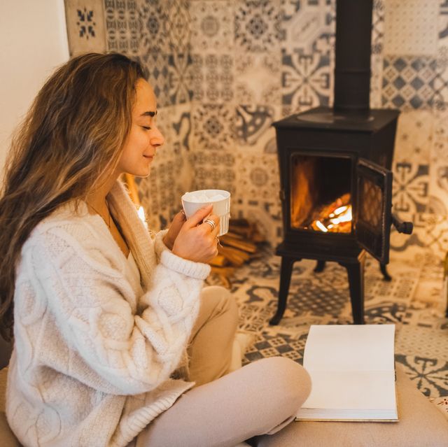 young woman drinking hot coffee and reading book on pillows near fireplace christmas eve