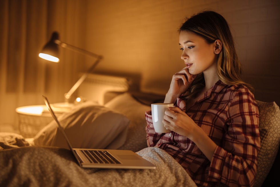 young woman holding a mug with her hot drink, while lying on her bed with laptop