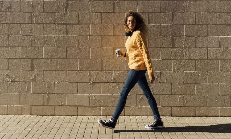 young woman holding disposable cup while walking on footpath by brick wall during sunny day