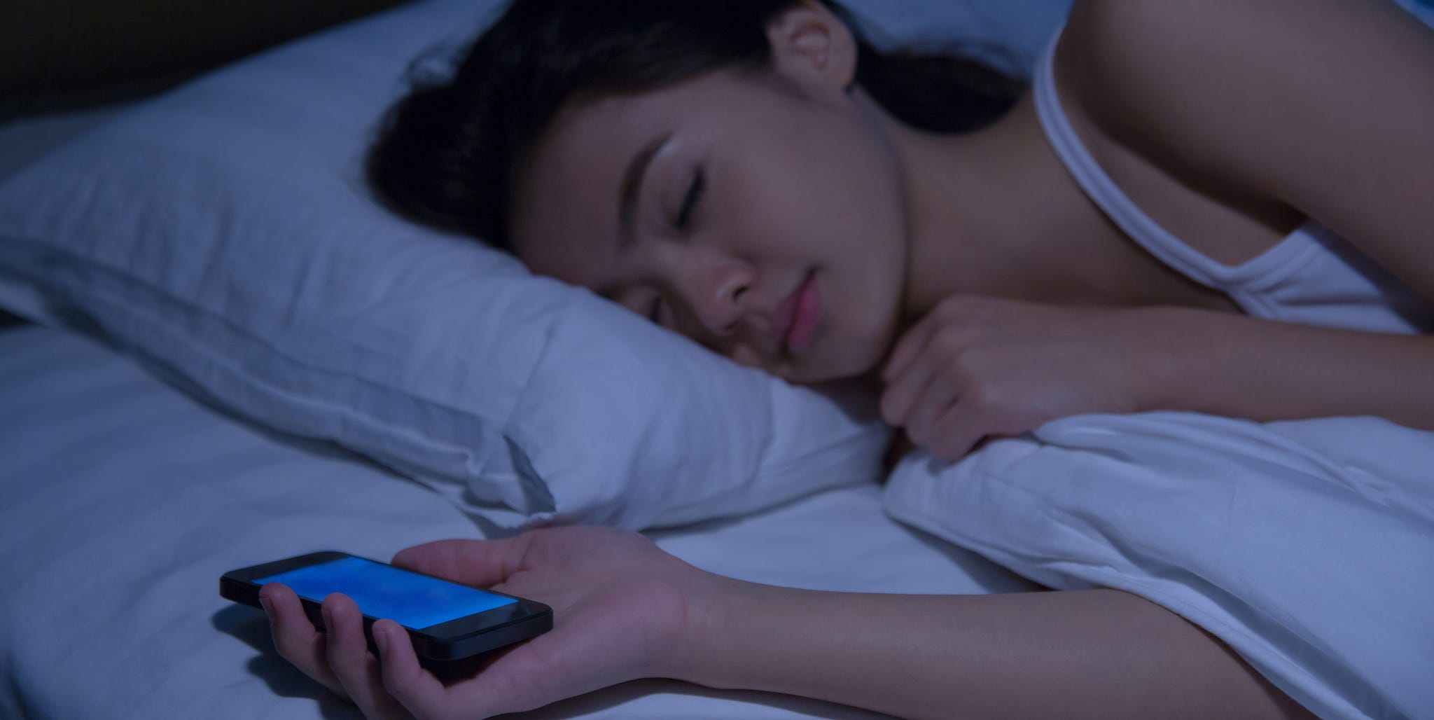 Young Woman Holding Mobile Phone While Sleeping On Bed At Home